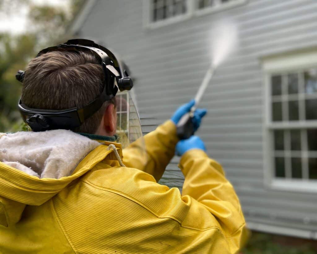 Professional wearing protective gear pressure washing the exterior siding of a house and showing how pressure washing works.